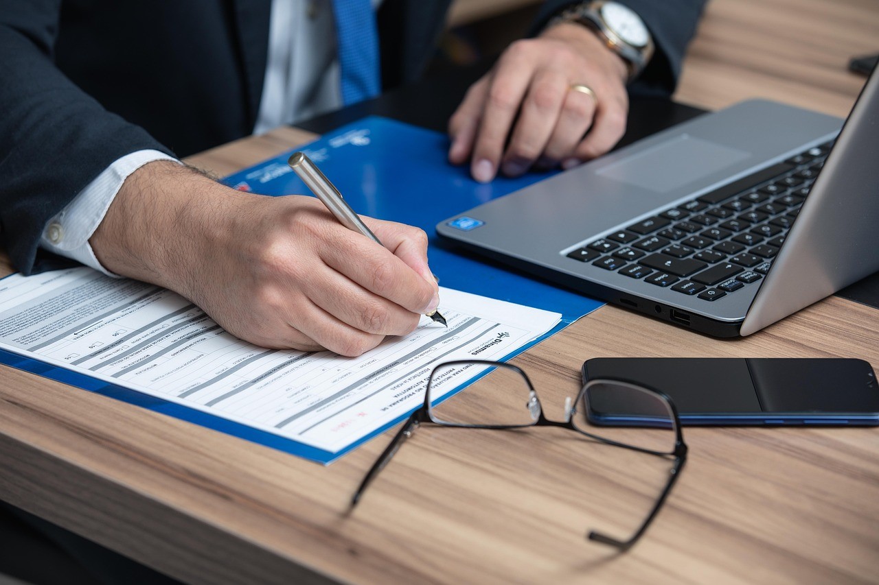 Close-up of a lawyer's hands filling out a form on a desk next to a laptop, smartphone, and a pair of glasses